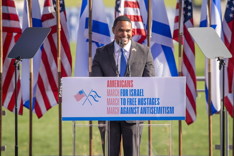 Representative Ritchie Torres speaks at a lectern that reads "Americans March for Israel March to Free Hostages March Against Antisemitism." Behind him are a row of U.S. and Israeli flags.