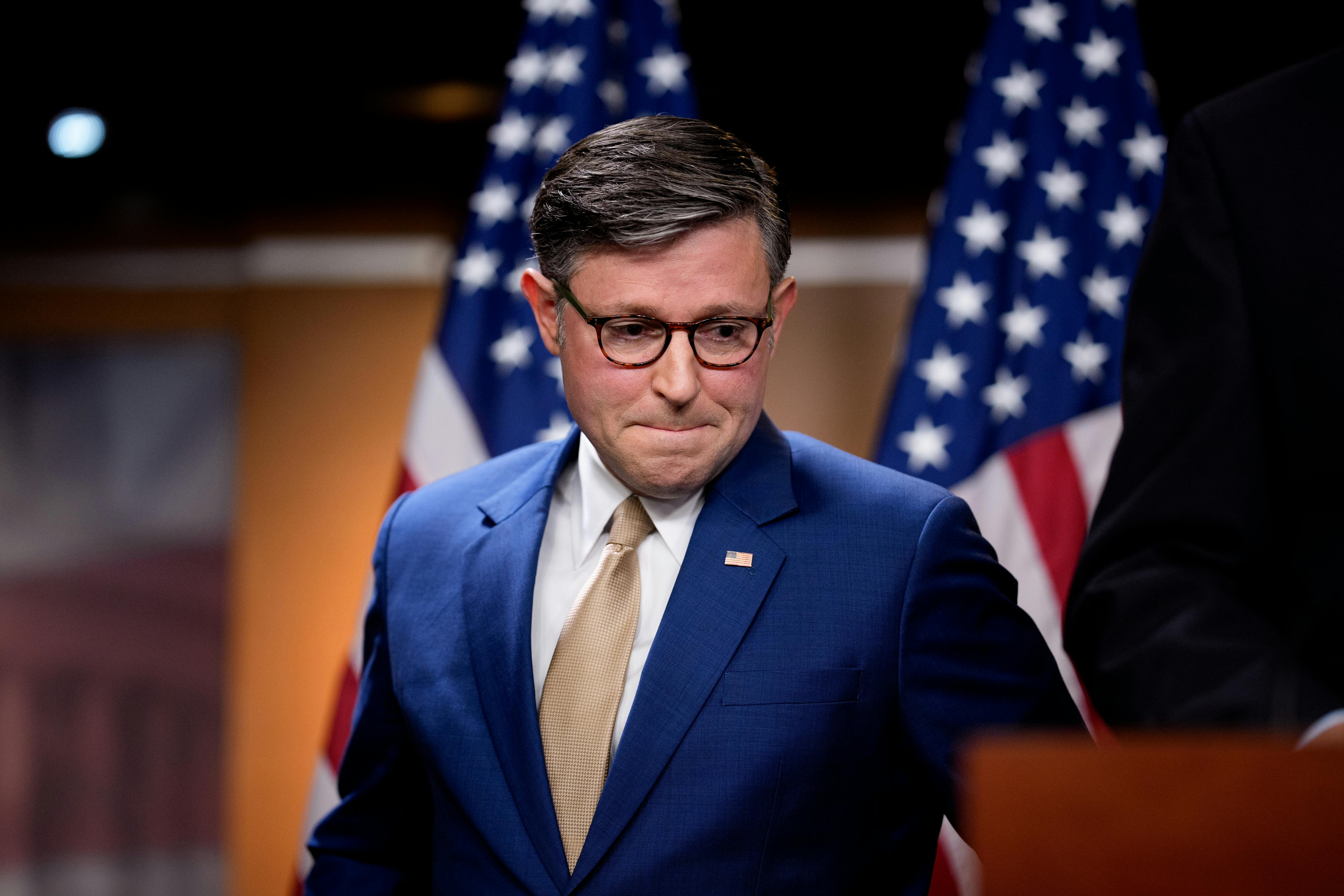 House Speaker Mike Johnson presses his lips together and looks down while standing next to a podium