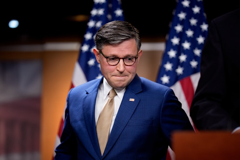 House Speaker Mike Johnson presses his lips together and looks down while standing next to a podium