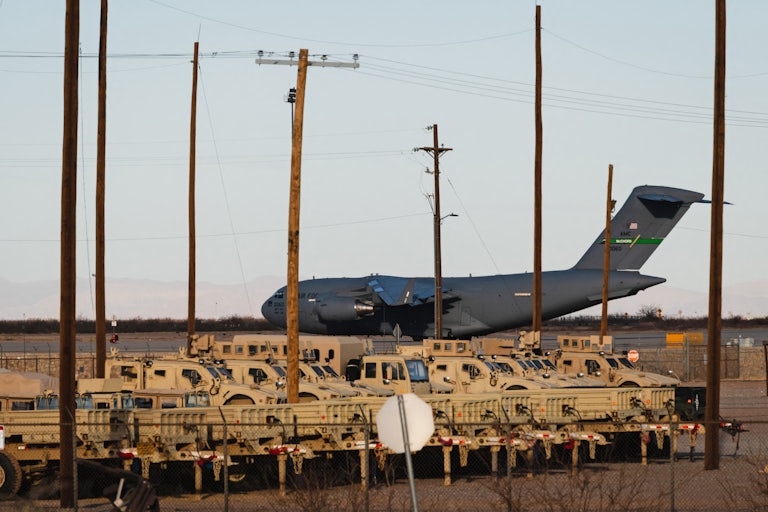 A plane sits on a runway in front of several military videos
