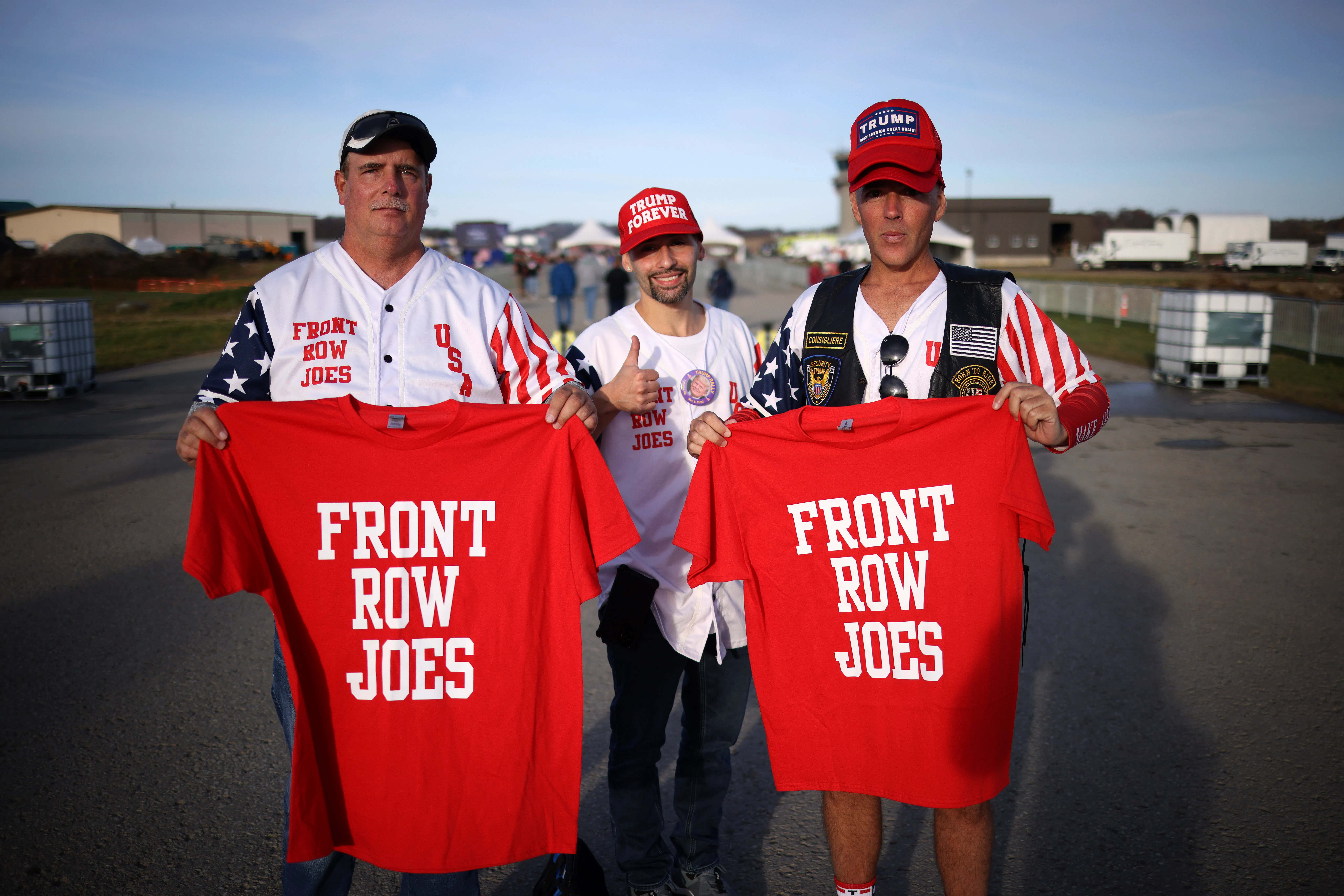 Two guys hold up red shirts that say "Front Row Joes" while a third wearing a "Trump Forever" cap gives a thumbs up