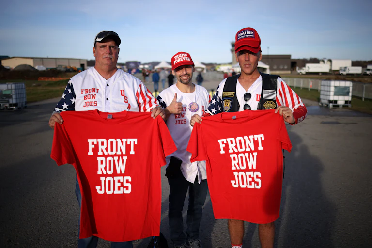 Two guys hold up red shirts that say "Front Row Joes" while a third wearing a "Trump Forever" cap gives a thumbs up