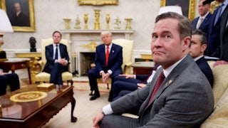 National Security Adviser Mike Waltz frowns at a camera while seated in the White House. Donald Trump can be seen in the background.