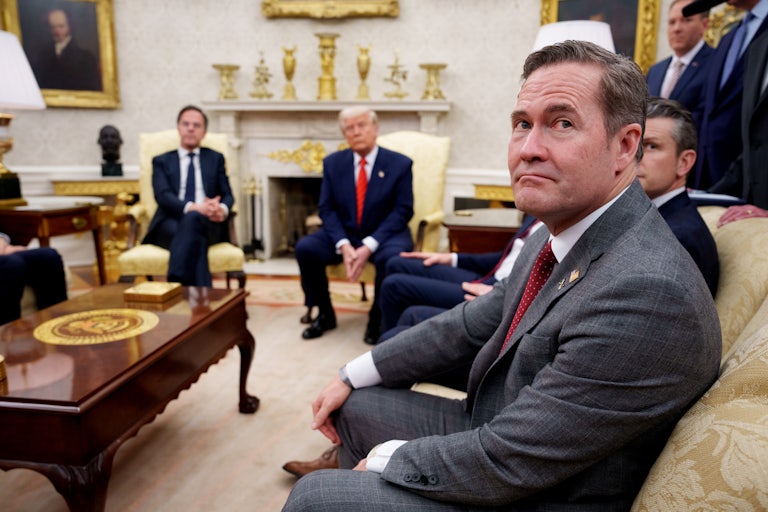 National Security Adviser Mike Waltz frowns at a camera while seated in the White House. Donald Trump can be seen in the background.