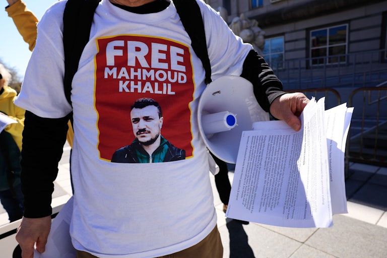 A person wears a shirt that says, "Free Mahmoud Khalil" holds a bullhorn and a stack of papers