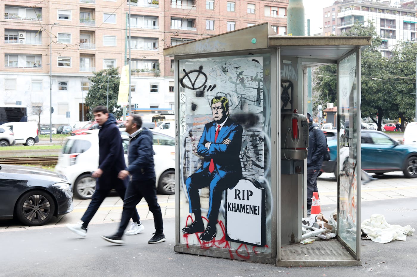 A photograph of people walking past a bus stop that has a mural of President Donald Trump seated with his arms crossed atop a tombstone inscribed RIP Khamenei/