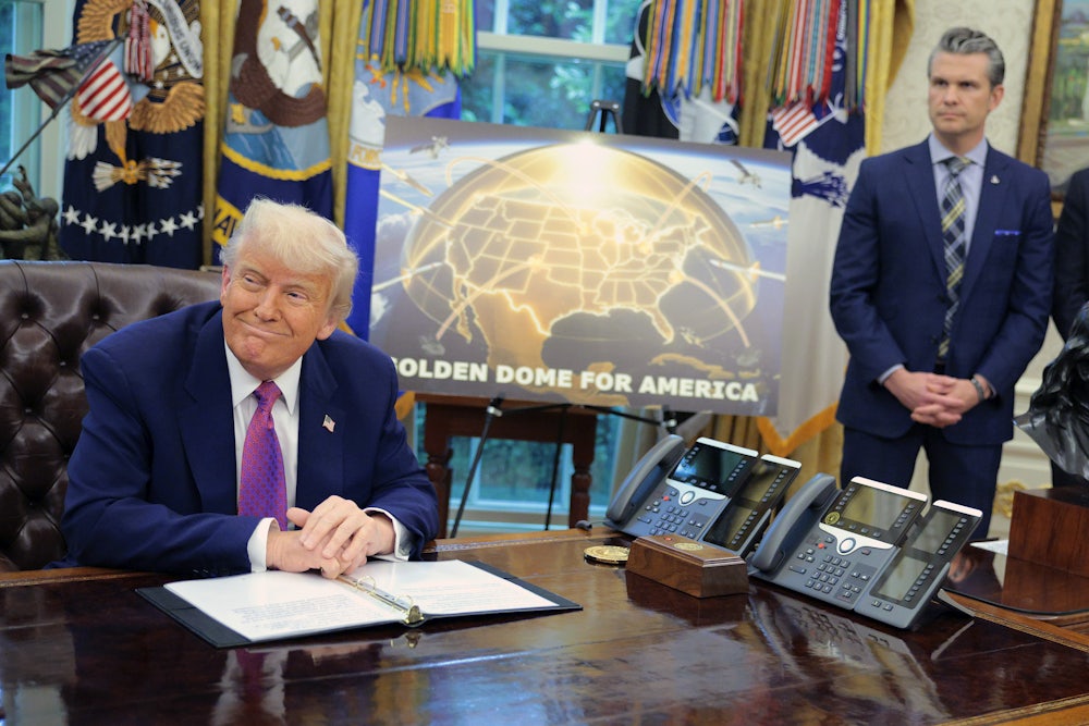 Donald Trump sits at his desk in the Oval Office of the White House. Behind him is a sign that displays a mock "Golden Dome." Defense Secretary Pete Hegseth stands next to the sign with his hands clasped in front of him.
