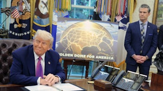 Donald Trump sits at his desk in the Oval Office of the White House. Behind him is a sign that displays a mock "Golden Dome." Defense Secretary Pete Hegseth stands next to the sign with his hands clasped in front of him.