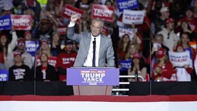 RFK Jr raises his fist while standing at a podium, with fans behind him.