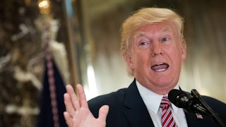 Trump stands at a microphone, holding a hand up, with his mouth open to talk.