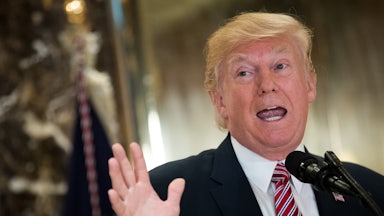 Trump stands at a microphone, holding a hand up, with his mouth open to talk.