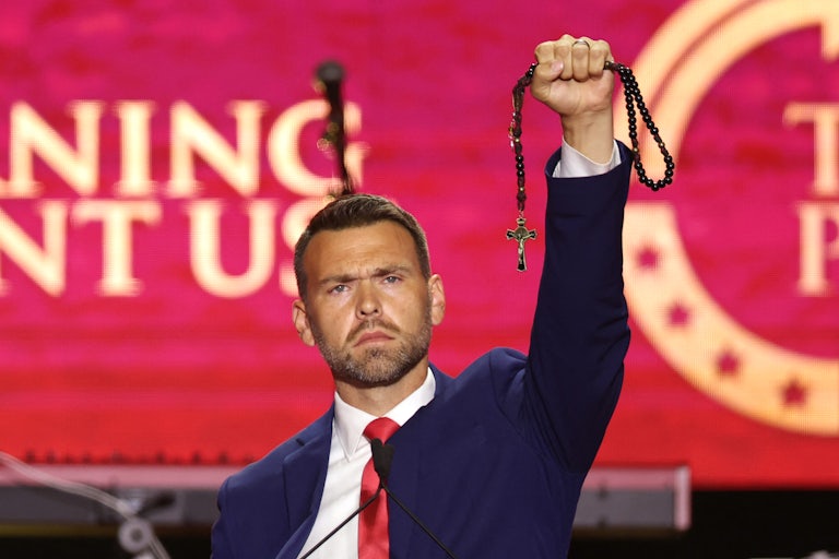 Jack Posobiec lifts a necklace with a cross on it while on the stage at a Turning Point conference.