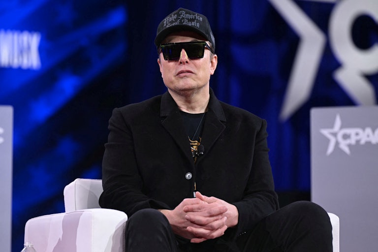 Elon Musk wears sunglasses indoors and a black MAGA hat.