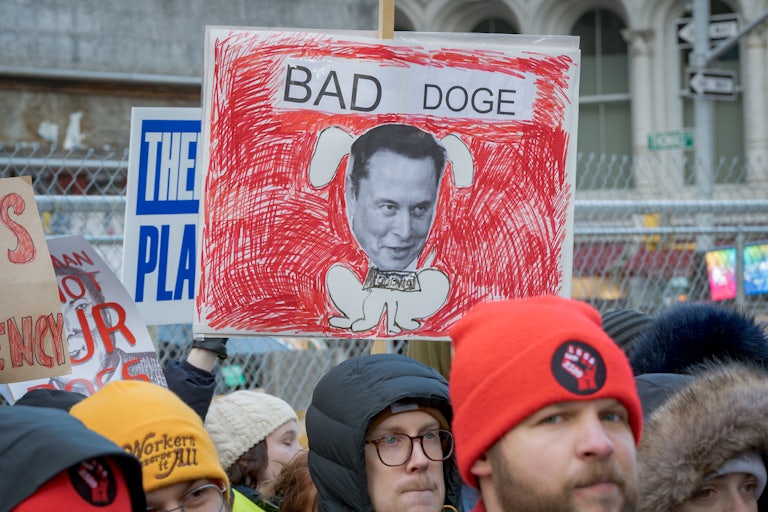 A protester holds up a drawing of Elon Musk as a dog with a dollar in its mouth. The words "Bad DOGE" are written above him