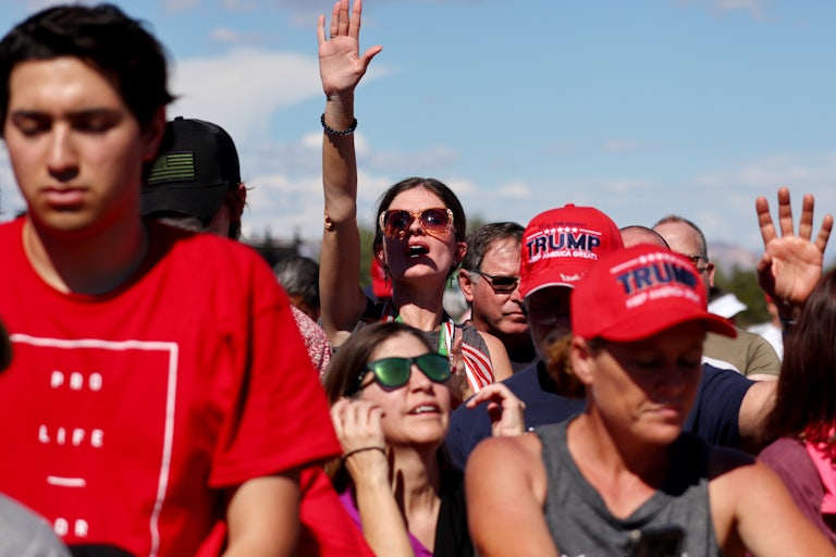 Several people stand in the sun wearing red shirts and red Trump caps. Two people raise their hands in the air.