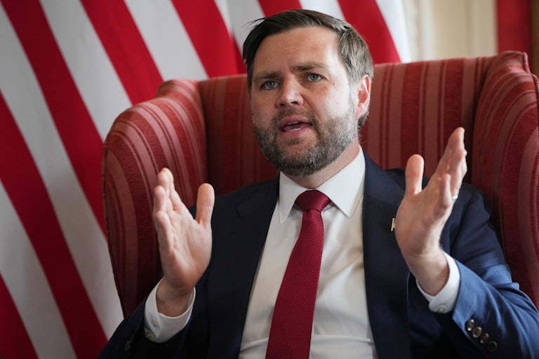 JD Vance sits on a chair and raises both hands while speaking