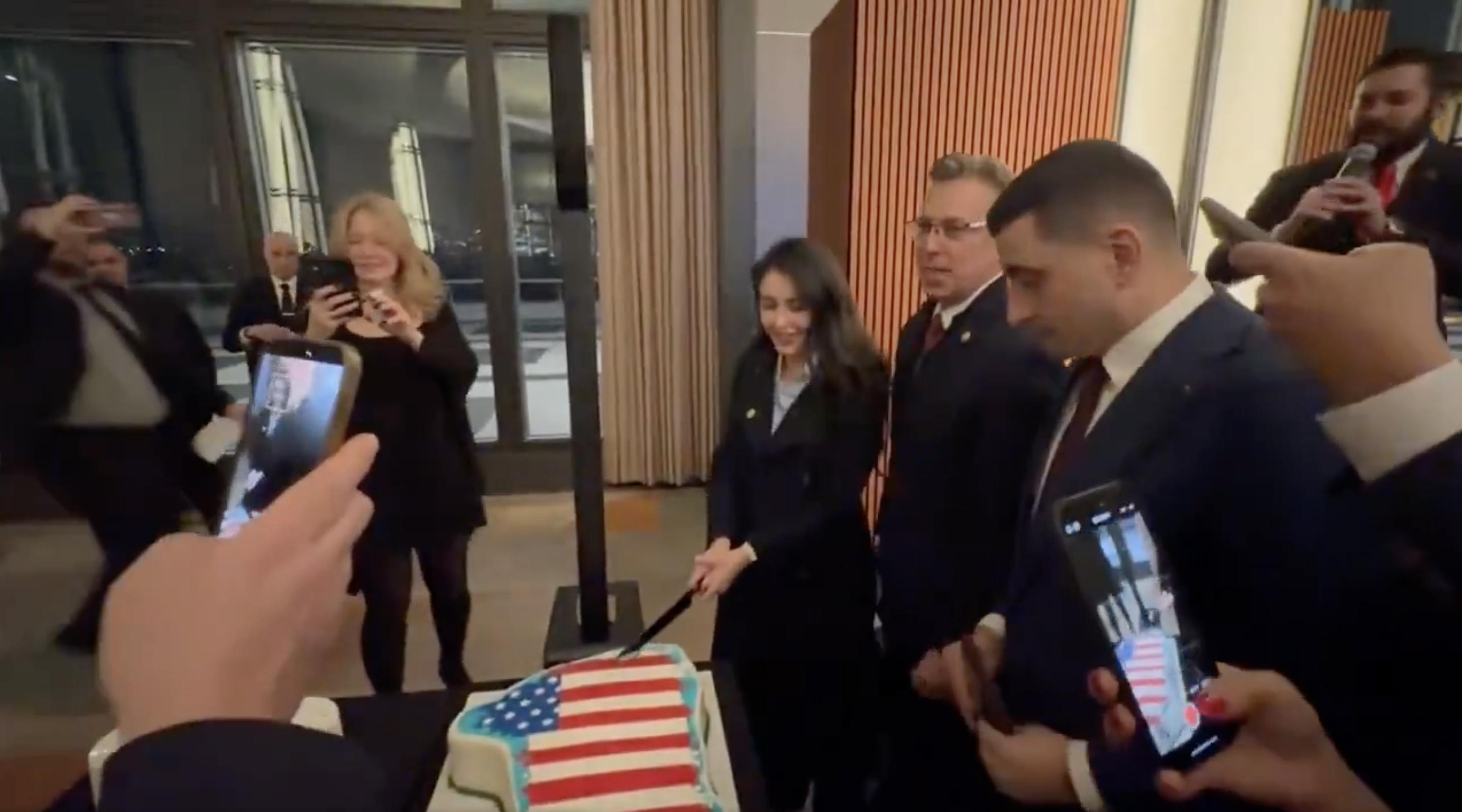 Representative Anna Paulina Luna cuts into a Greenland flag draped in the U.S. flag alongside Representative Andy Ogles and Romania's George Simion. Others hold up their cameras to record.