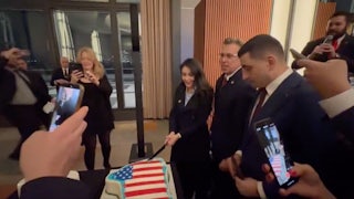 Representative Anna Paulina Luna cuts into a Greenland flag draped in the U.S. flag alongside Representative Andy Ogles and Romania's George Simion. Others hold up their cameras to record.