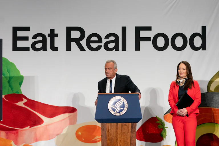Robert F. Kennedy Jr. leans forward slightly and speaks at a podium in front of a banner that says, "Eat Real Food." Agriculture Secretary Brooke Rollins stands next to him.