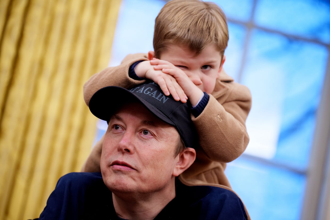 Elon Musk's toddler son sits on his shoulders and leans forward on his Make America Great Again cap.