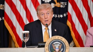 President Donald Trump speaks into a mic while seated behind the presidential seal.