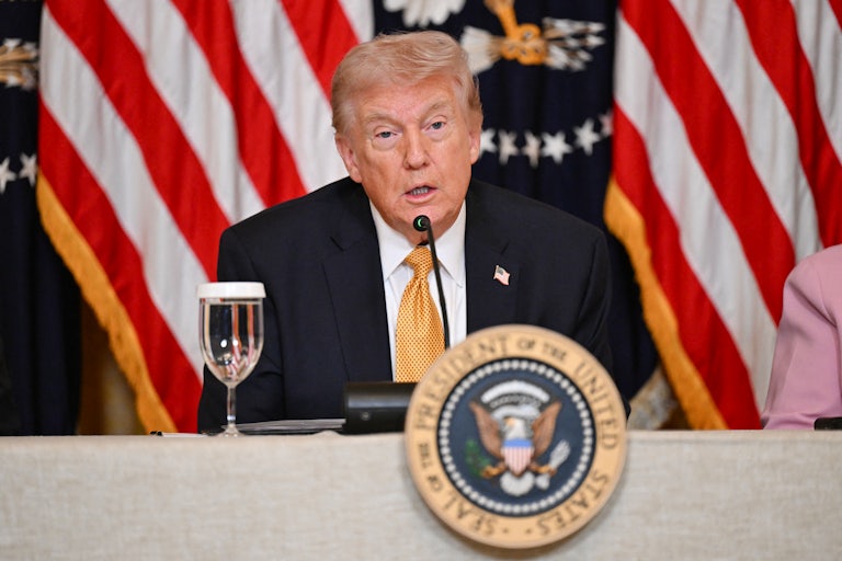 President Donald Trump speaks into a mic while seated behind the presidential seal.