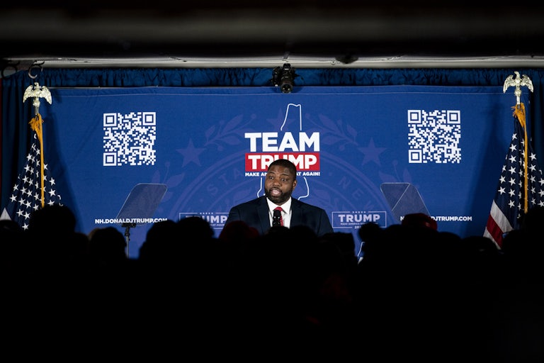 Byron Donalds stands on a stage in front of a background that says "Team Trump" and has a QR code. A crowd's silhouette is seen in front of him.
