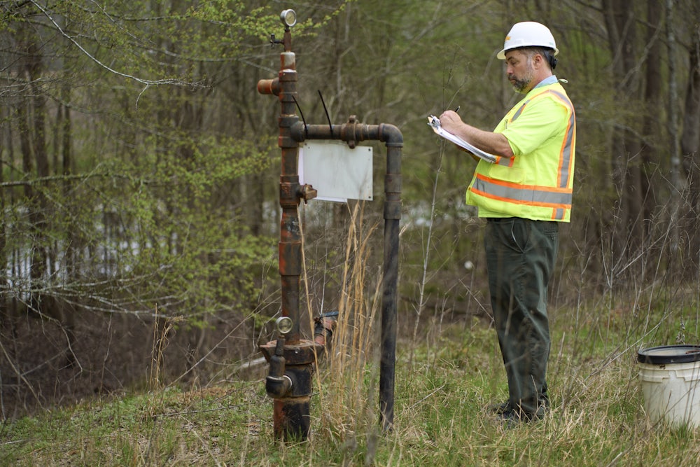 A person in a reflective jersey writes on a clipboard next to an orphaned well.