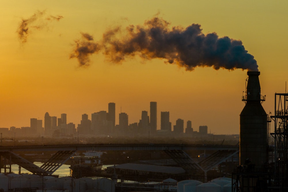 A smokestack churns out smoke and vapor with a city in the distance against an orange sky