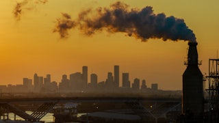 A smokestack churns out smoke and vapor with a city in the distance against an orange sky