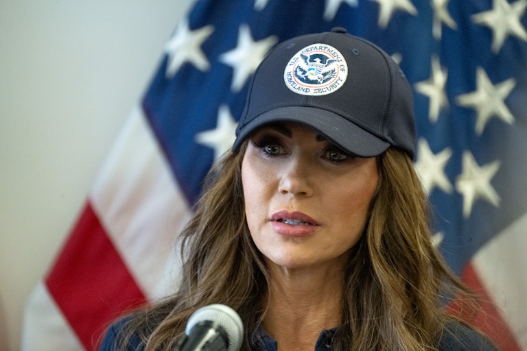 Homeland Security Secretary Kristi Noem wears a DHS cap and speaks into a mic.