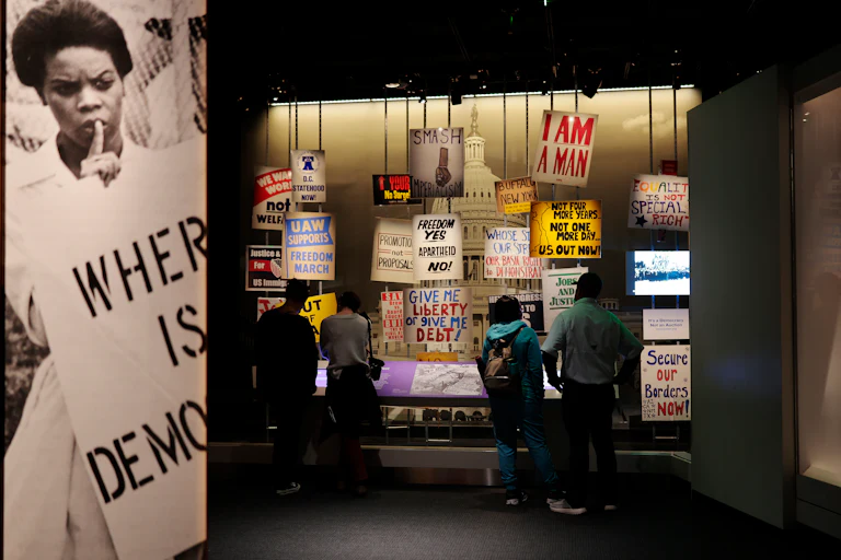 An exhibit at the National Museum of American History features signs like "Where is democracy," "Freedom Yes Apartheid No," and "Give me liberty or give me debt."
