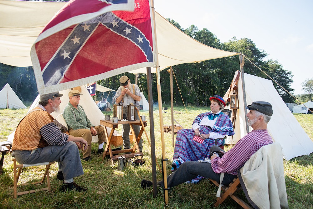 A photo of Confederate reenactors at a campsite wearing Civil war clothing with a Confederate flag flying nearby.