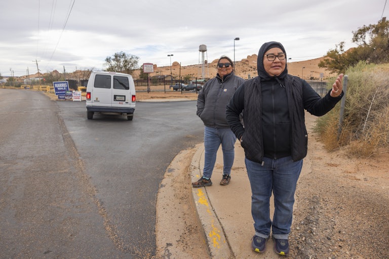 Two Native women stand on the sidewalk as a white fan is seen in the background