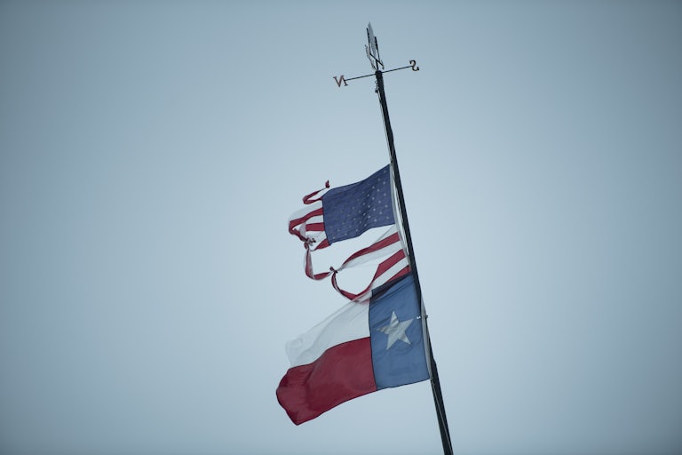 The state flag of Texas hangs on a staff below a tattered American flag.