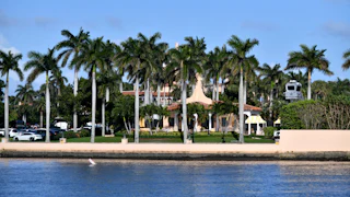 Donald Trump's Mar-a-Lago mansion is visible behind water and palm trees, with a parking lot to its right.