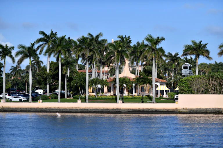 Donald Trump's Mar-a-Lago mansion is visible behind water and palm trees, with a parking lot to its right.