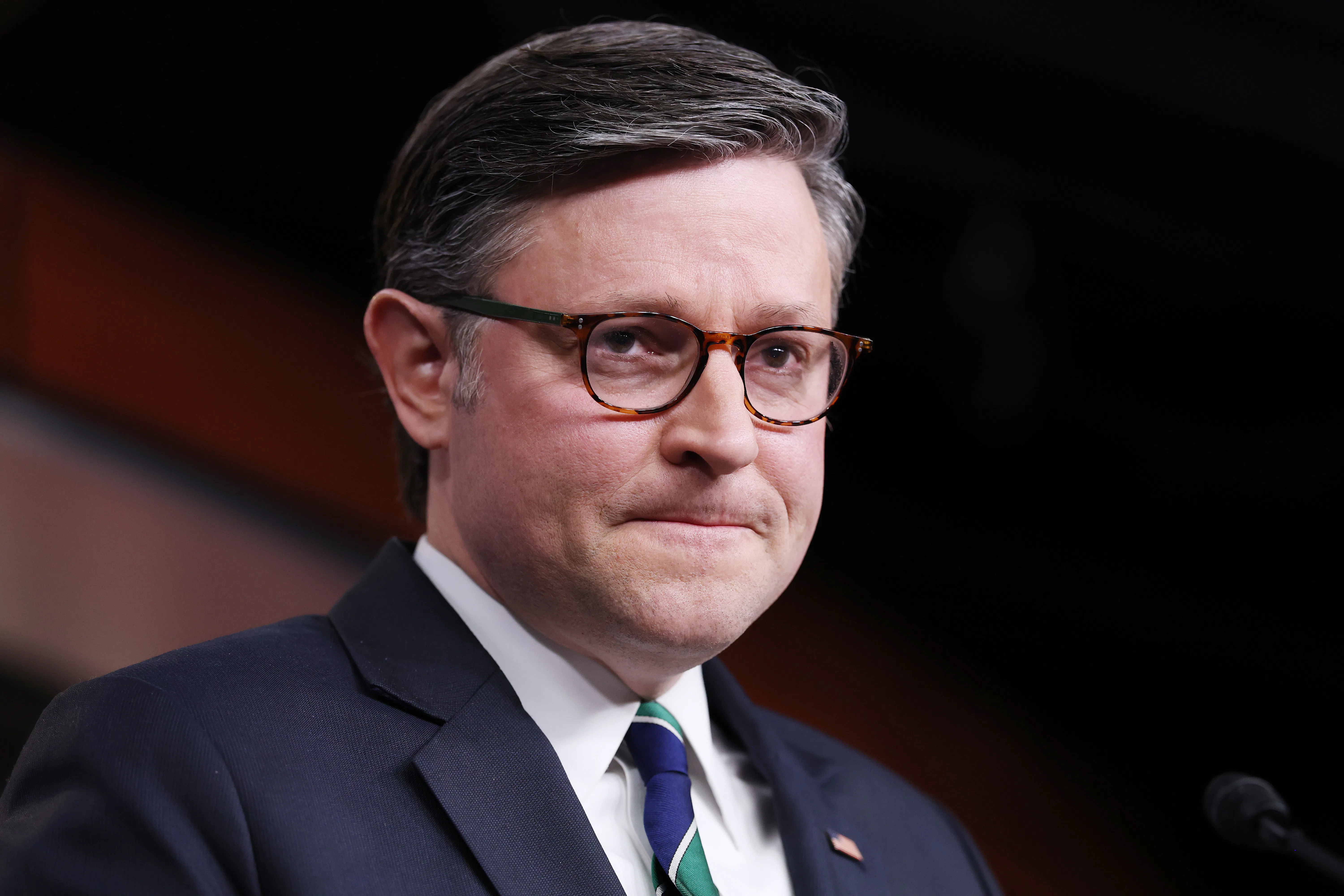 Mike Johnson viewed at an angle wearing a blue suit and striped tie with a frustrated expression on his face where his lips are pressed closely together.