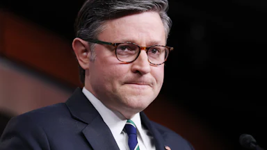 Mike Johnson viewed at an angle wearing a blue suit and striped tie with a frustrated expression on his face where his lips are pressed closely together.