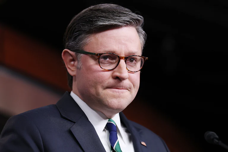 Mike Johnson viewed at an angle wearing a blue suit and striped tie with a frustrated expression on his face where his lips are pressed closely together.