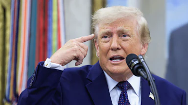Donald Trump points his right finger at his head while grinning