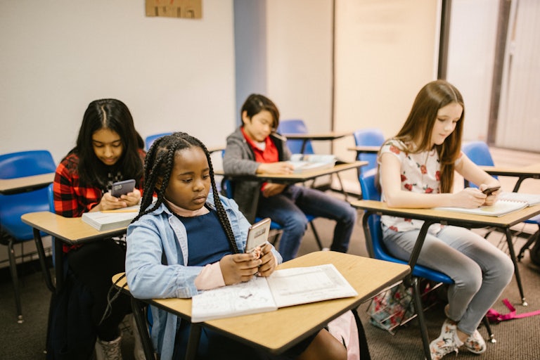 Three young girls sit at their desks, looking down at their phones.