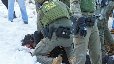 Border Patrol agents (faces unpictured) slam someone into the snow, face-down.