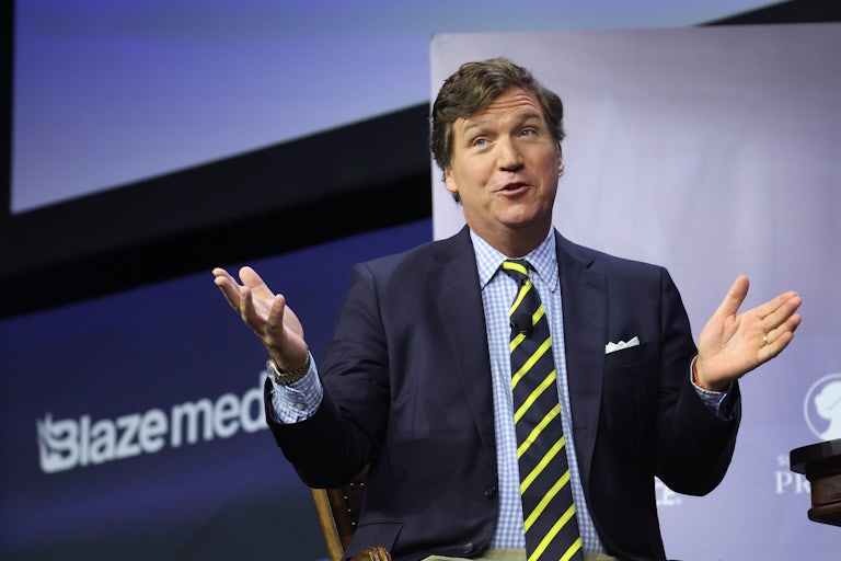 Tucker Carlson is speaking on stage and raises his hands in the air as if to shrug