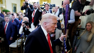 Donald Trump walks with two baseball bats in the White House as reporters take photos of him.