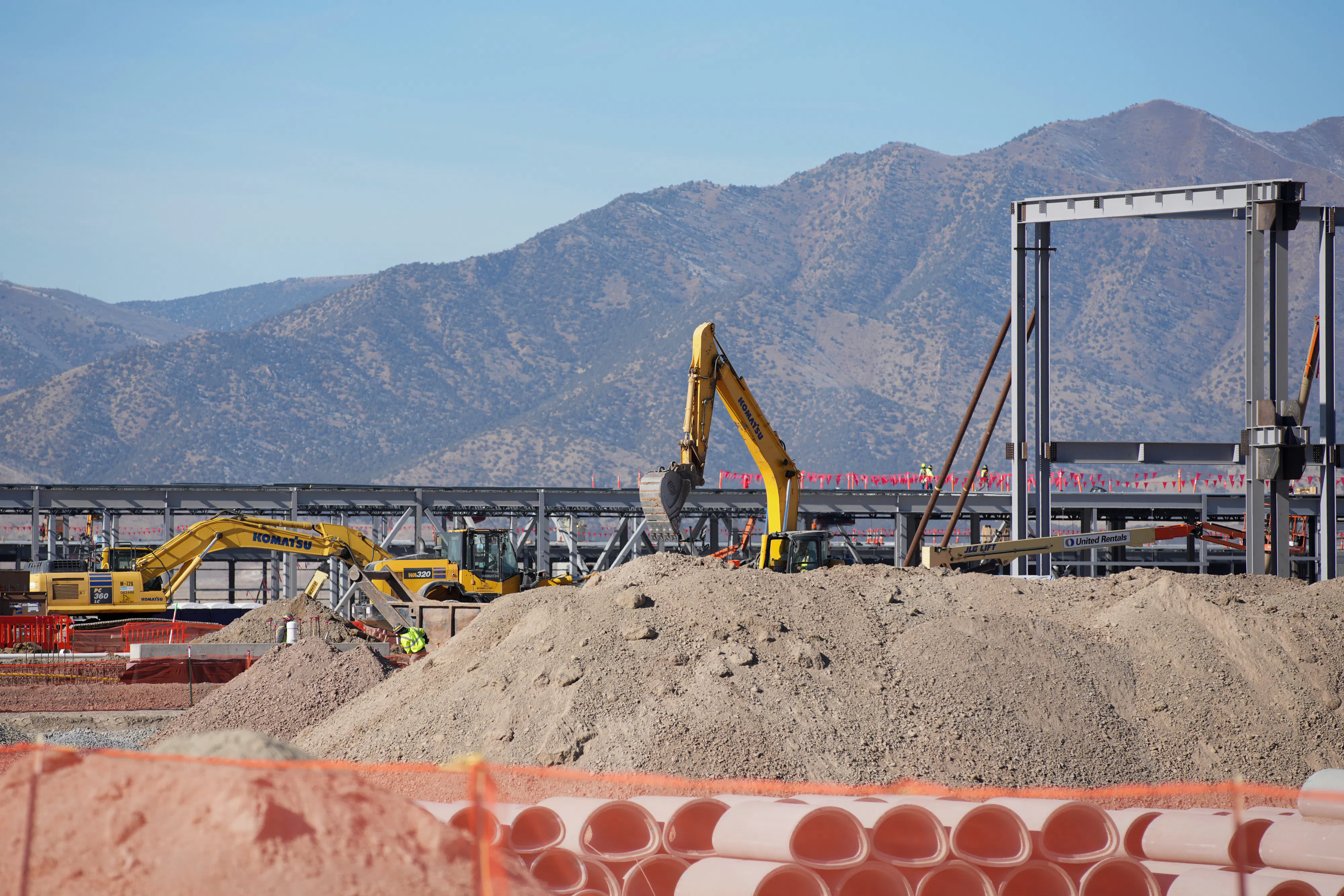 An excavator stands amid construction equipment and piles of dirt, with mountains in the background.