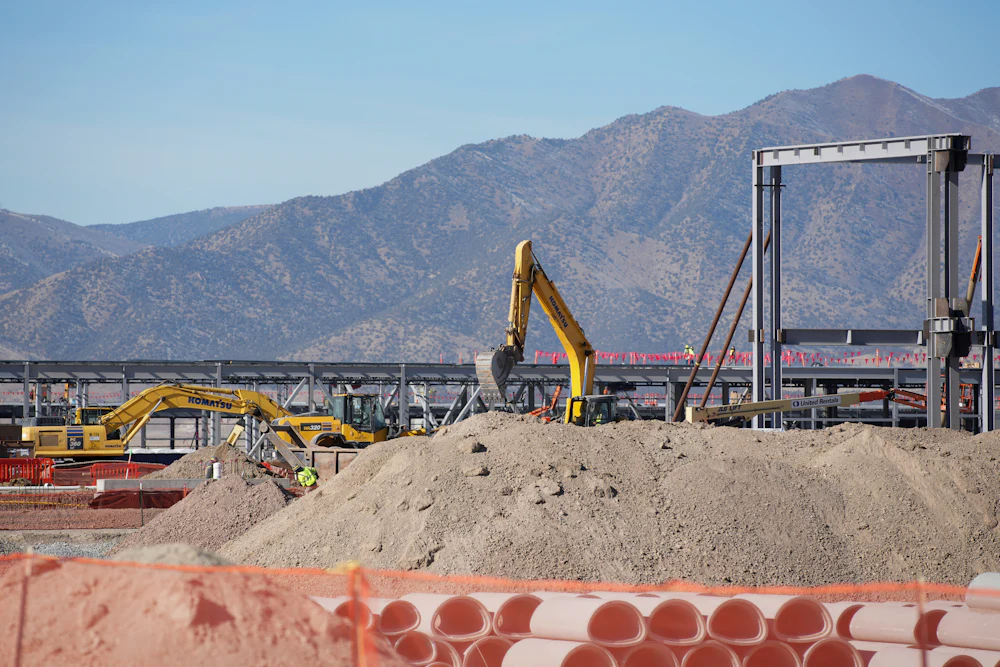 An excavator stands amid construction equipment and piles of dirt, with mountains in the background.