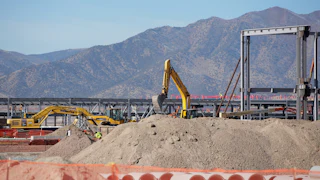 An excavator stands amid construction equipment and piles of dirt, with mountains in the background.