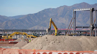An excavator stands amid construction equipment and piles of dirt, with mountains in the background.