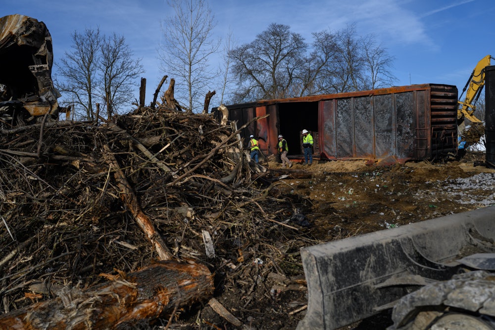 A cargo train, a giant pile of branches/uprooted trees, and three workers in the background.